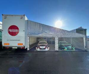 An image of two Raceway Motorsport cars next to the transport vehicle that has the Prosura logo on the back.