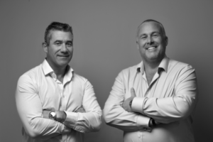 The image shows Paul Schofield (on the left) and Jon Newall (on the right) in a black and white photo, both smiling with their arms crossed.