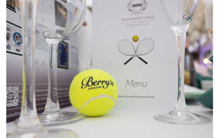 Image of a Berry's Tennis Event branded tennis ball placed on a table with champagne flutes and a wine list.