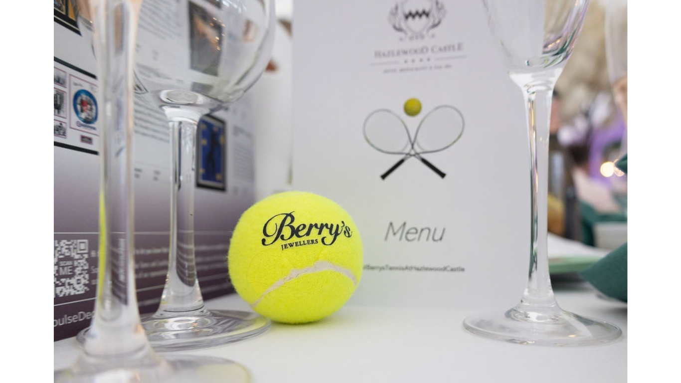 Image of a Berry's Tennis Event branded tennis ball placed on a table with champagne flutes and a wine list.